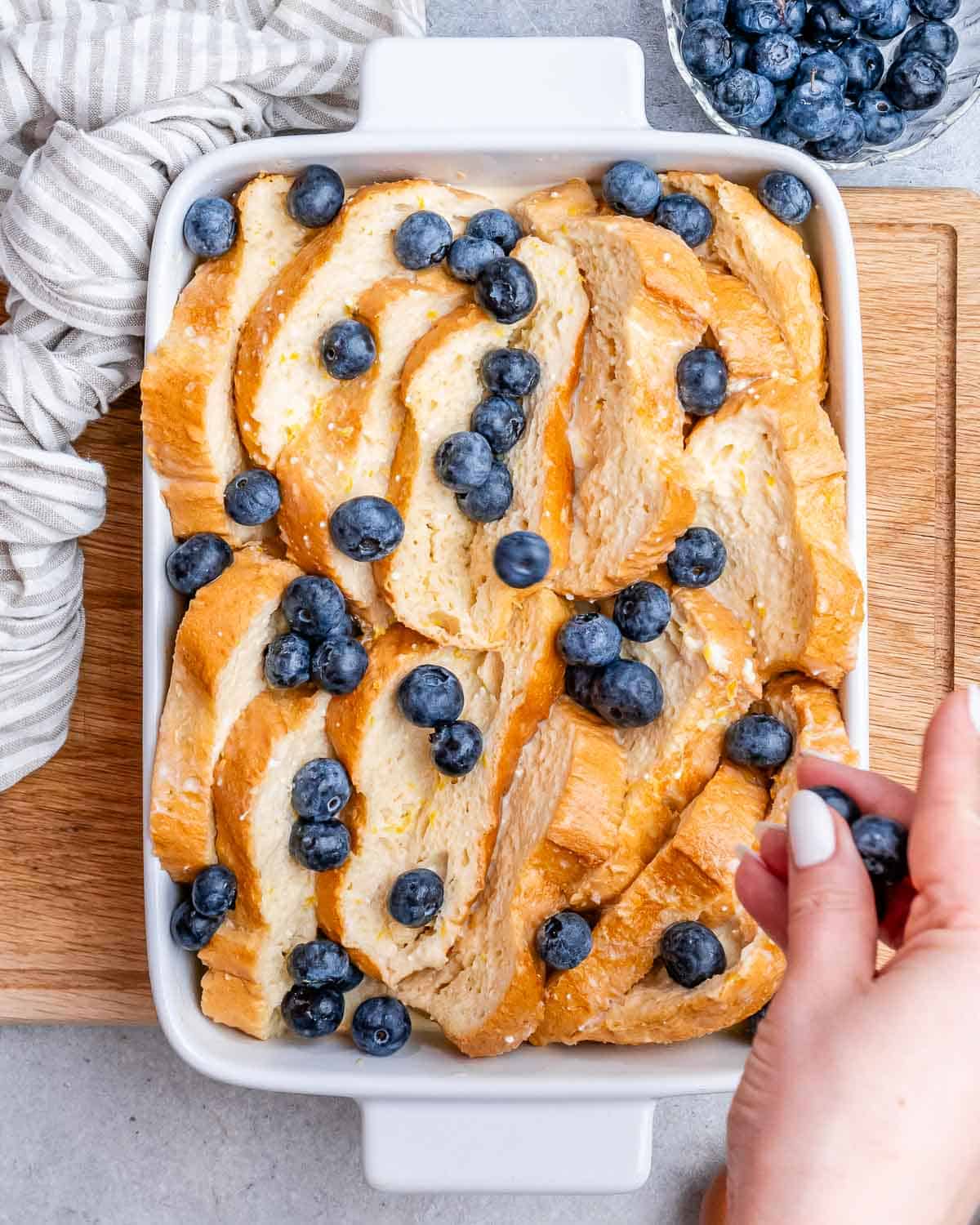 A hand placing blueberries on uncooked French toast bake.