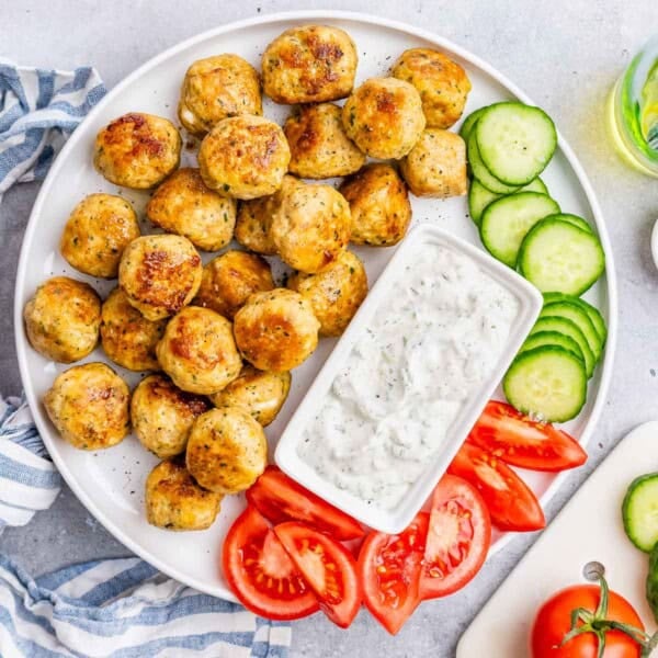 Meatballs on a plate with fresh cut up cucumbers, tomatoes, and a bowl of white sauce for dipping.