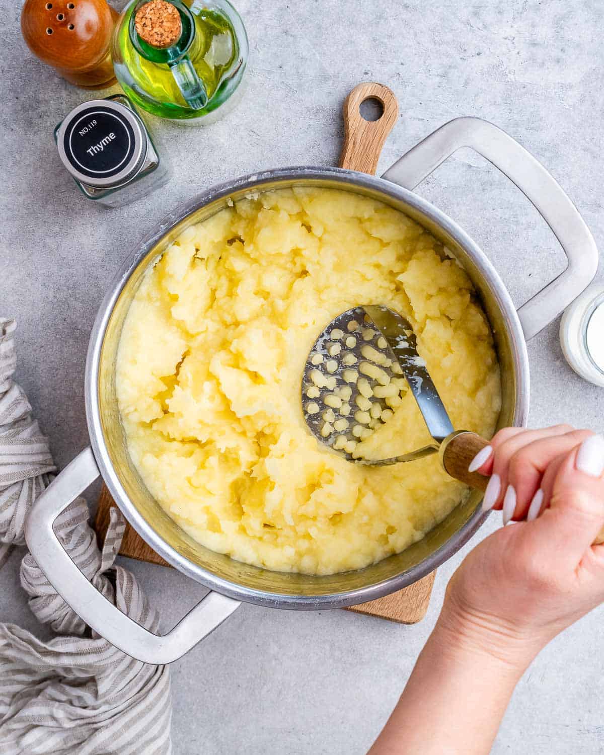 A hand using a potato masher to mash potatoes in a steel pot.