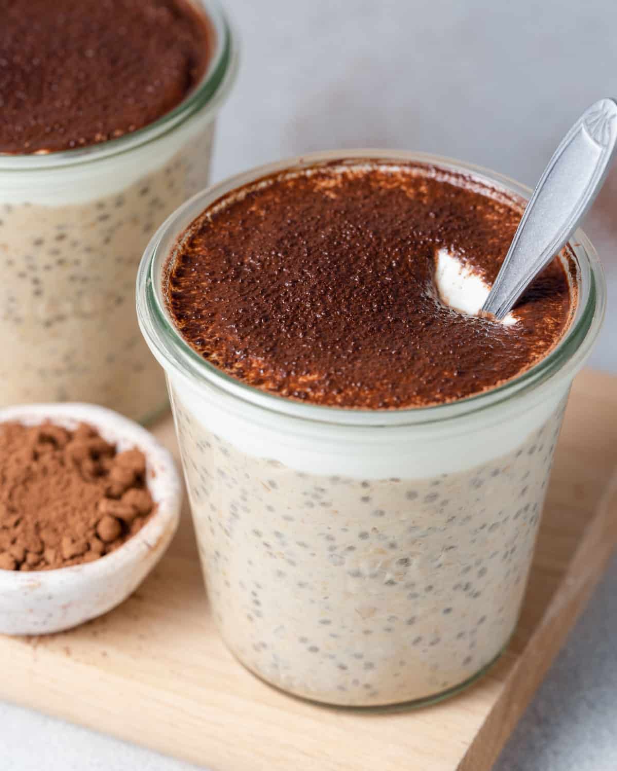 a jar on a cutting board filled with overnight oats, topped with cocoa powder, and spoon in the jar.