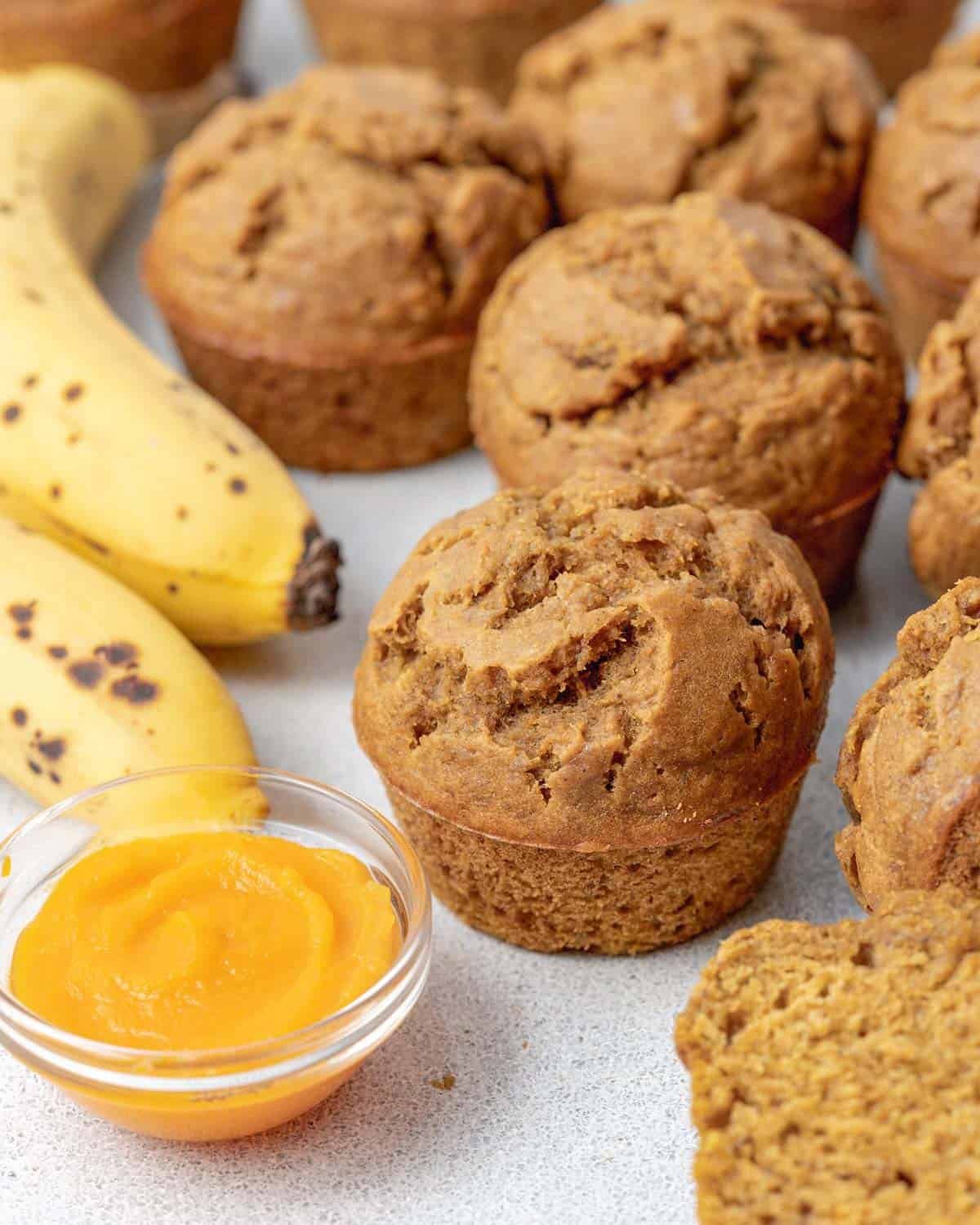 Pumpkin banana muffins on a white surface next to a bowl of pumpkin puree and some bananas.