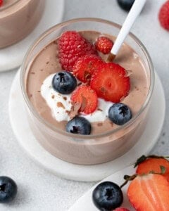 Close up of mousse in a glass cup with a spoon, topped with berries and set on a small white plate.