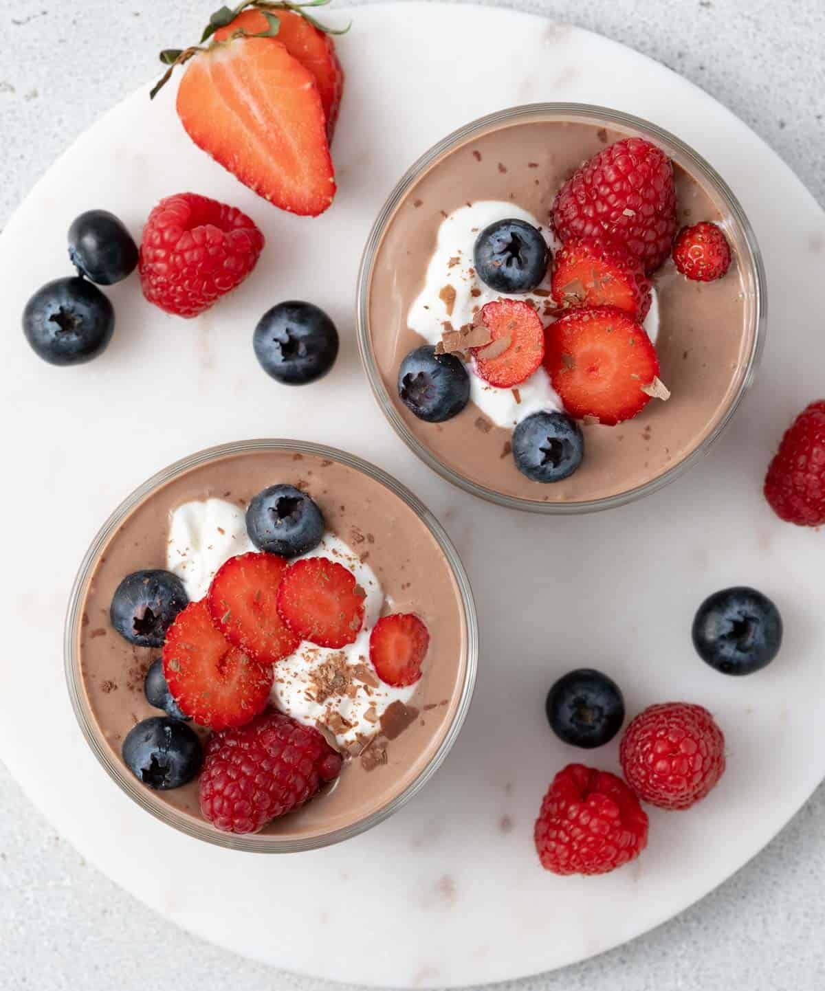 Top view of chocolate mousse topped with whipped cottage cheese, sliced strawberries, raspberries, and blueberries in glass cups on a white surface.