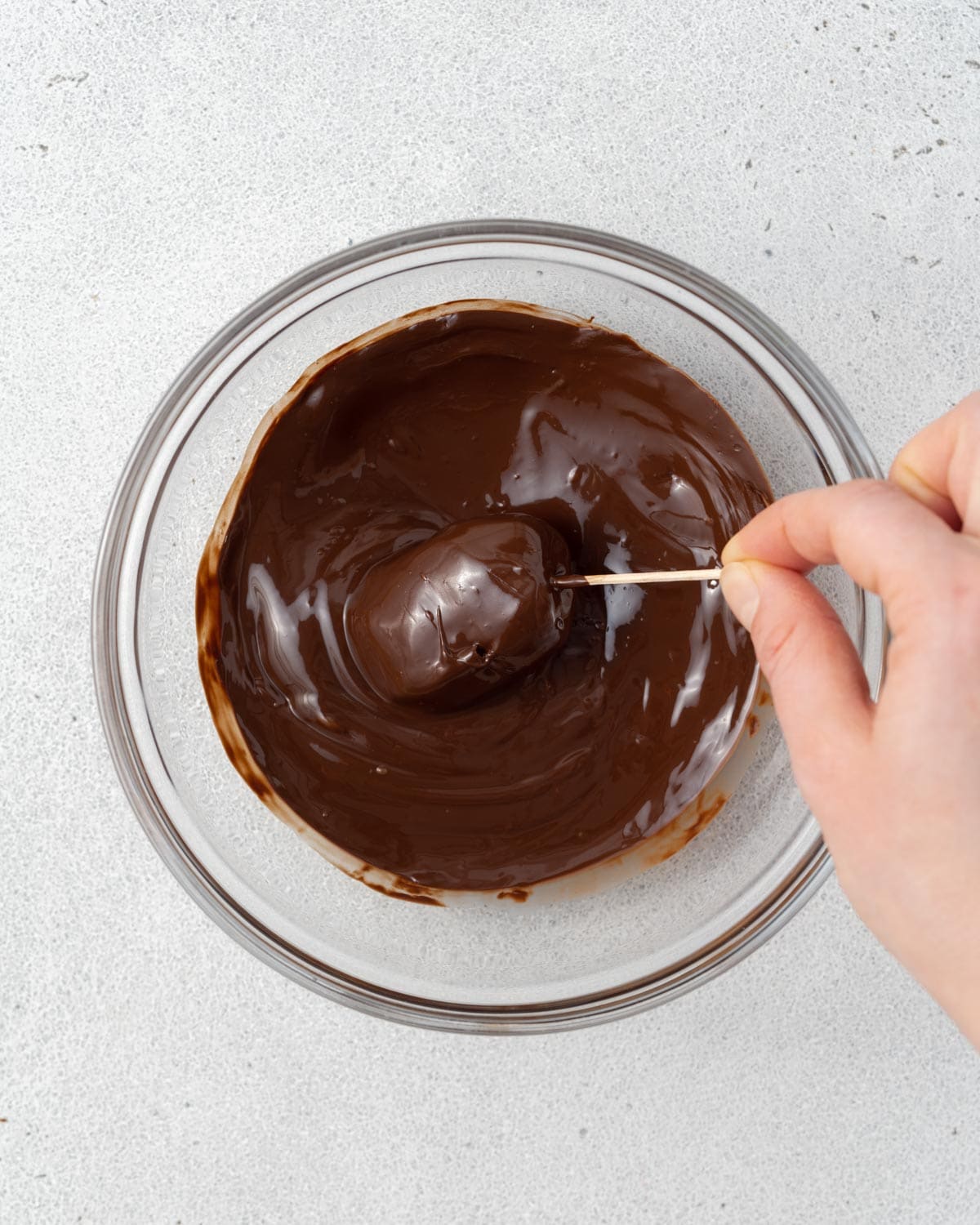 A hand using a toothpick to pierce the date and bring it out of the melted chocolate.