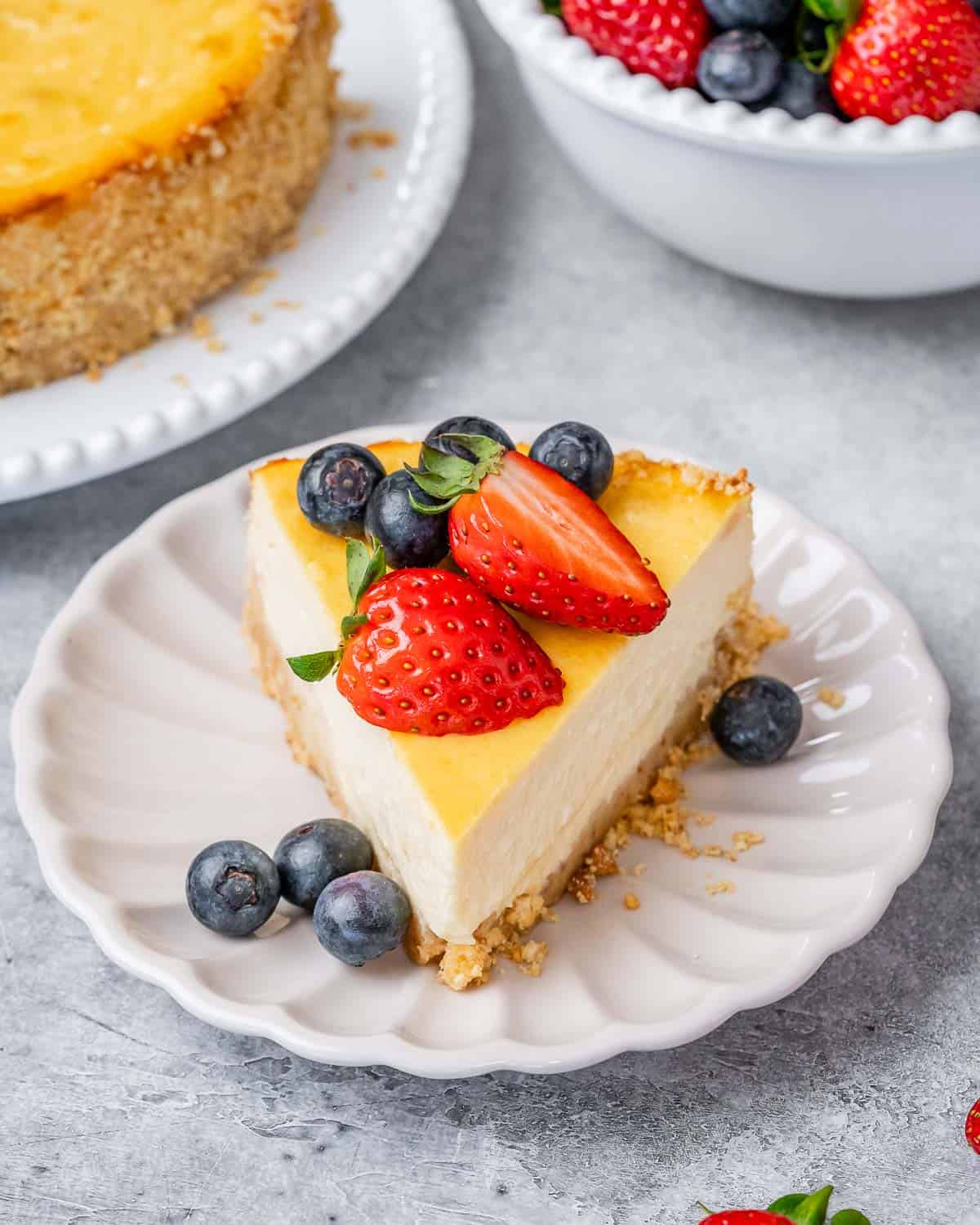 Cheesecake slice on a scalloped plate with fresh blueberries and halved strawberries.