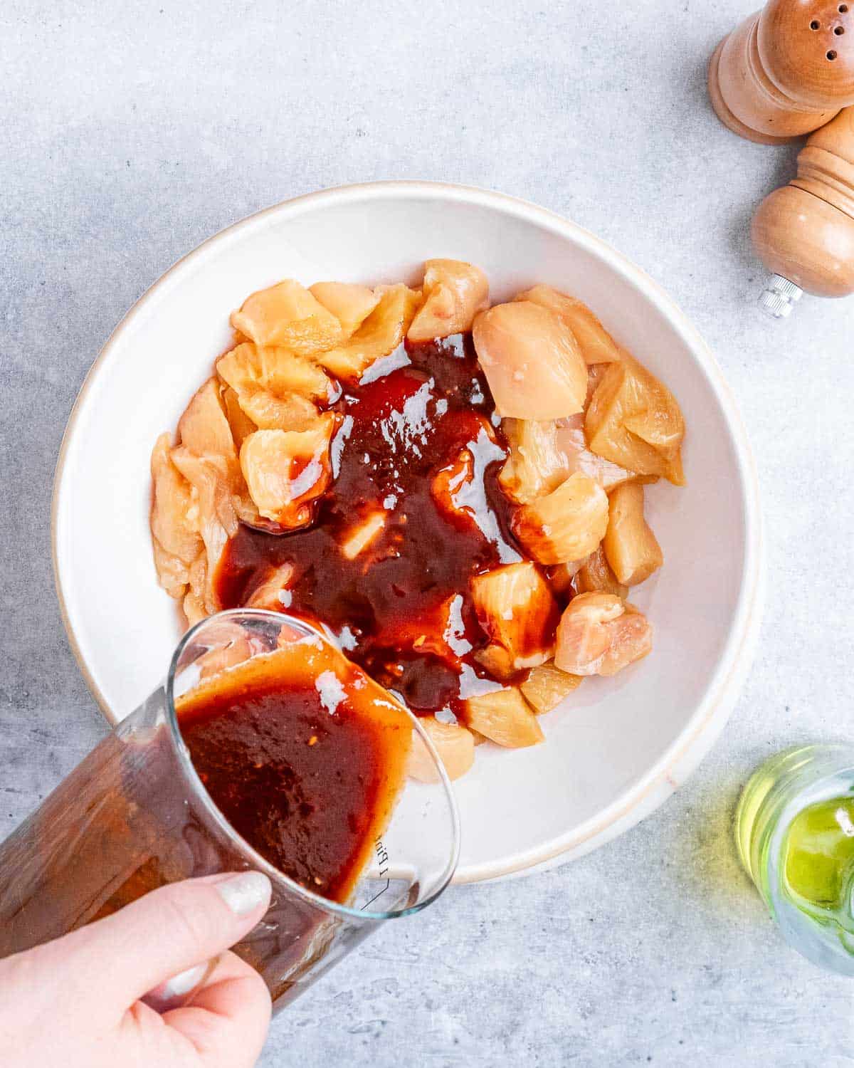 Pouring red sauce over raw chicken pieces in a white bowl.