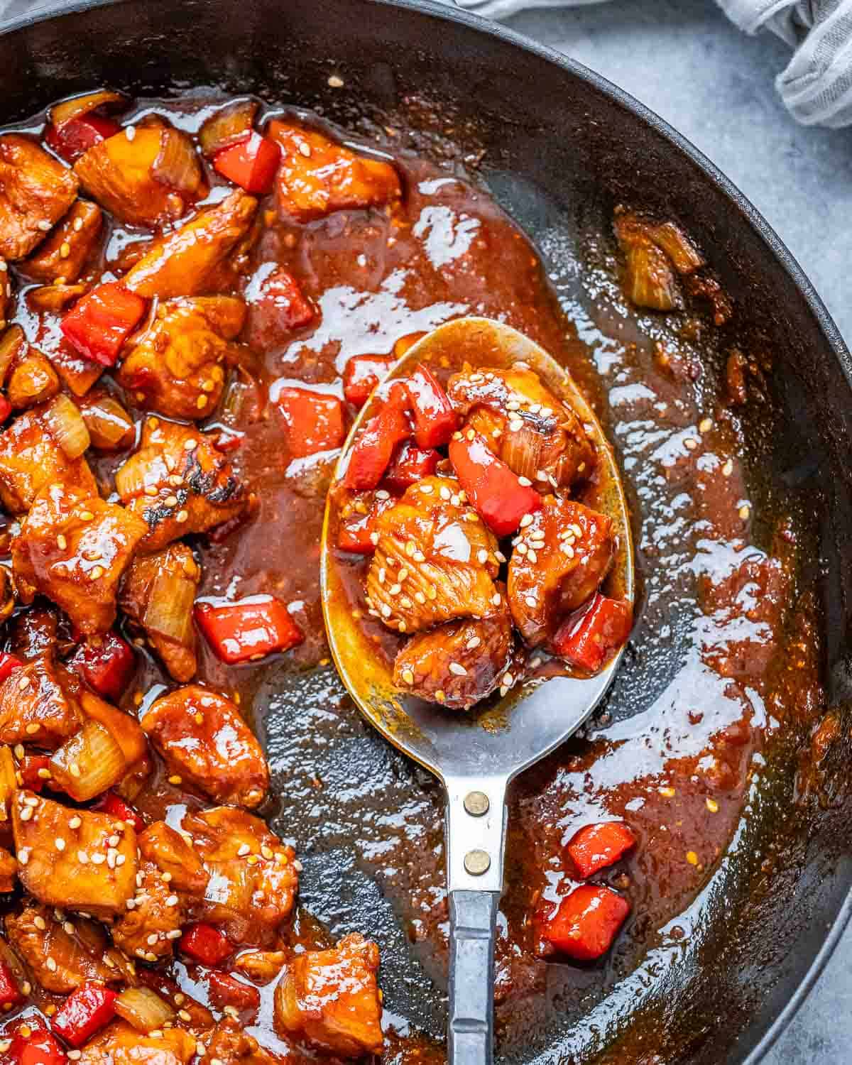 A spoon holding cooked chicken bites in a red sauce over a skillet.