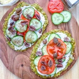 Two avocado flatbread on a cutting board topped with yogurt, tomatoes, cucumber, and olives.