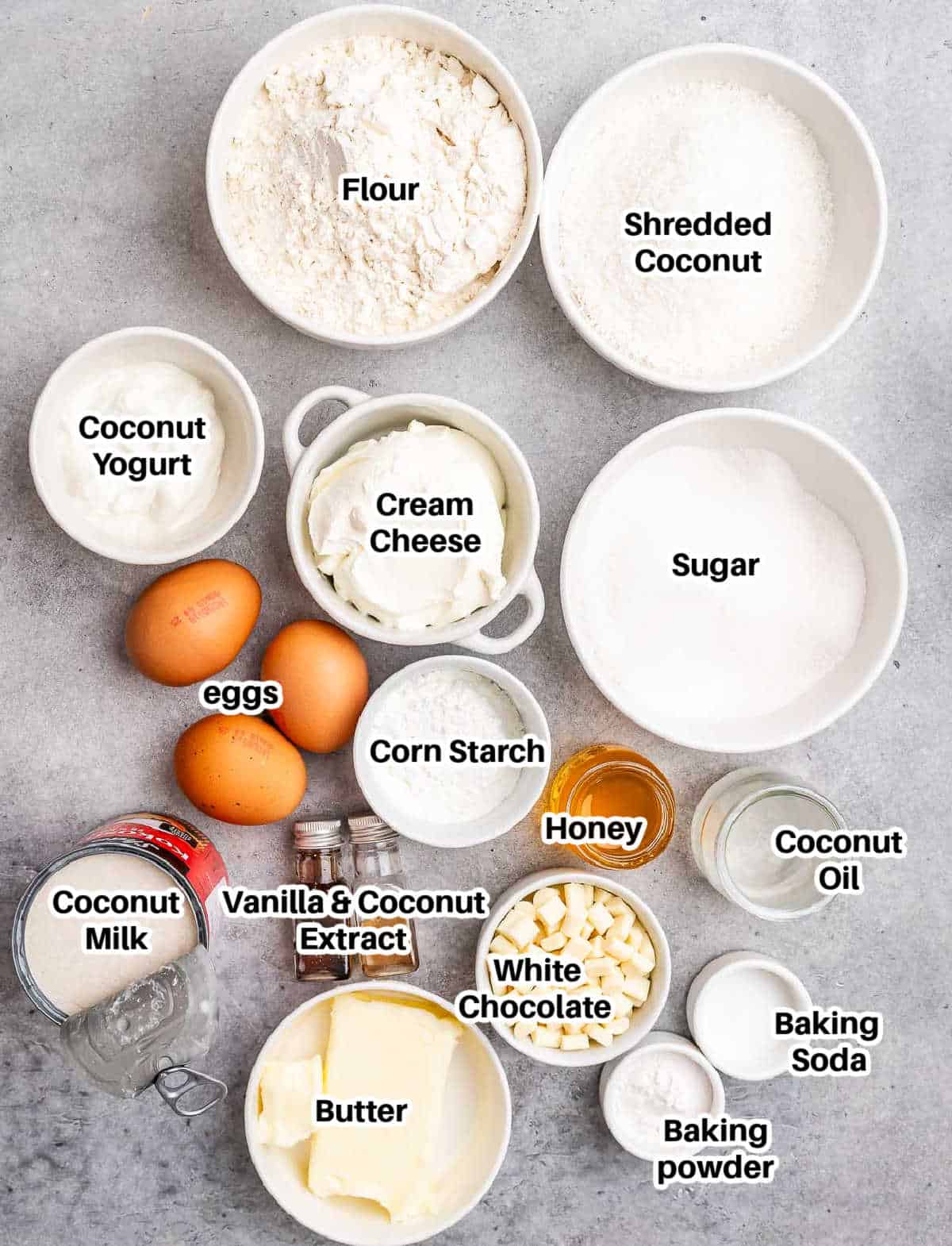 Overhead shot of labeled coconut cake ingredients including flour, eggs, coconut milk, shredded coconut, cream cheese, white chocolate chips, and others.