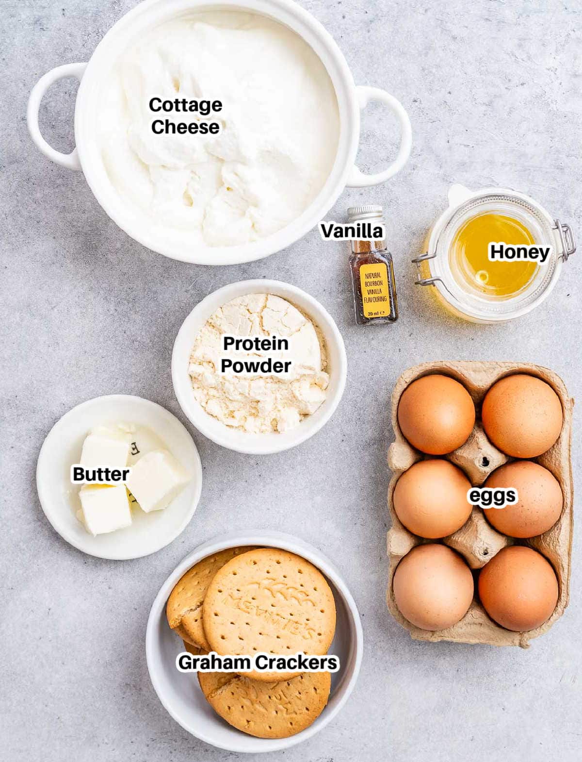 Top view of ingredients in small bowls on a white surface: cottage cheese, eggs, graham crackers, honey, protein powder, butter, and vanilla.