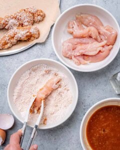 Raw chicken tenders next to bowls of buffalo sauce and breading station, ready to be dipped and coated.