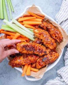 Fully cooked buffalo chicken tenders served in a bowl with fresh celery and carrot sticks, hand reaching in to grab one.