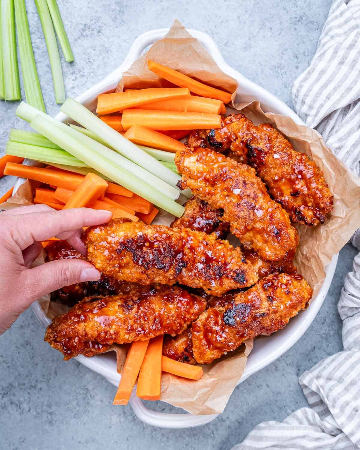 Fully cooked buffalo chicken tenders served in a bowl with fresh celery and carrot sticks, hand reaching in to grab one.