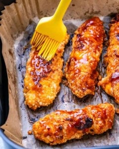 Crispy buffalo chicken tenders on parchment paper being brushed with buffalo sauce using a yellow silicone brush.