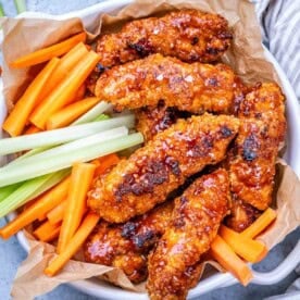 Pile of buffalo chicken tenders on parchment paper, served with carrot and celery sticks in a rustic basket.