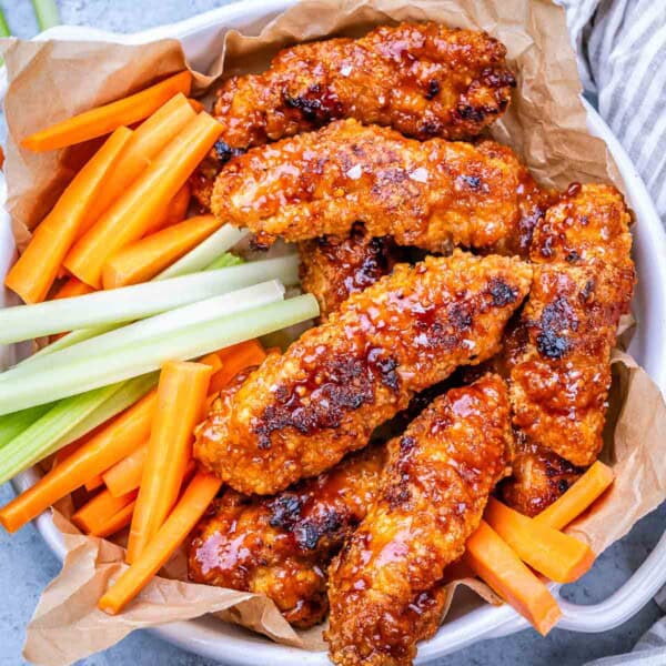 Pile of buffalo chicken tenders on parchment paper, served with carrot and celery sticks in a rustic basket.