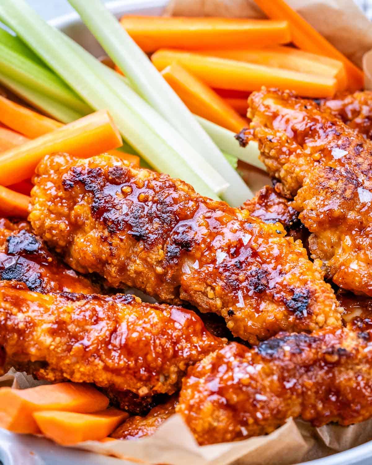 Close-up of crispy, saucy buffalo chicken tenders with celery and carrots, showing glossy texture and deep color.