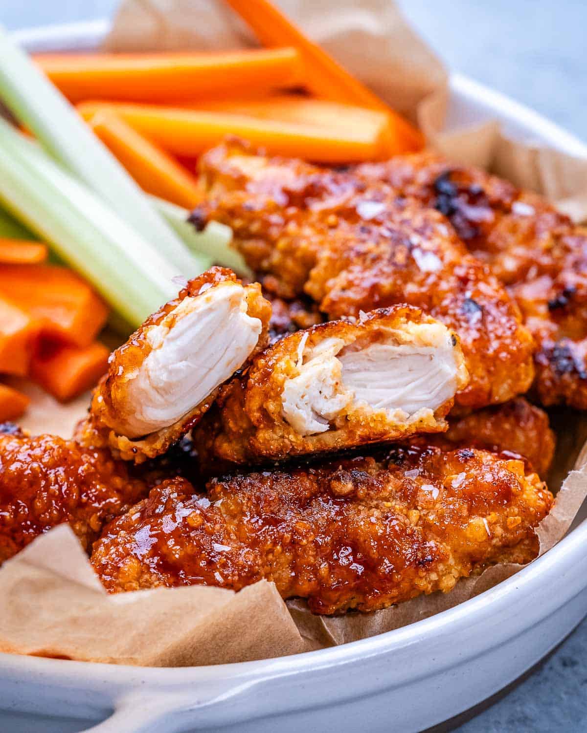 Cooked buffalo chicken tender with a bite taken out, showing juicy white interior, surrounded by celery and carrot sticks.