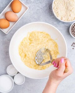 Mashed bananas being mashed with a masher in a white bowl, surrounded by baking ingredients.