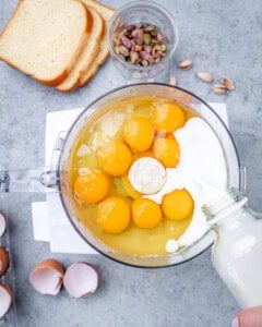 Food processor filled with blended pistachio mixture, with eggs and milk added on top before mixing into a custard.