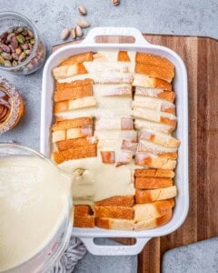 Pouring the pistachio custard over neatly layered brioche slices in a white casserole dish, preparing for baking.