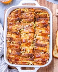 Baked Brioche French Toast Casserole in a white baking dish, golden brown on top with chopped pistachios scattered throughout.