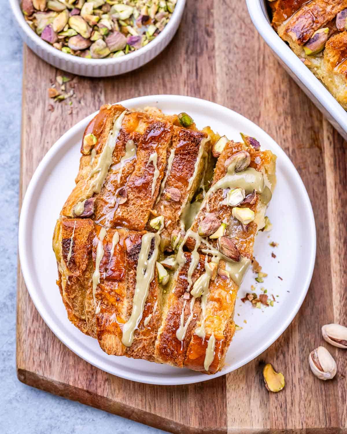 Slice of Brioche French Toast Casserole on a white plate, drizzled with pistachio butter and topped with crushed pistachios.