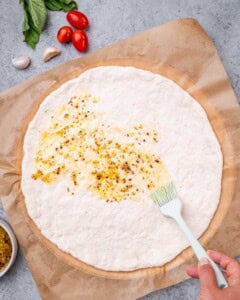 Raw pizza dough spread out on parchment paper with garlic oil being brushed over the top.