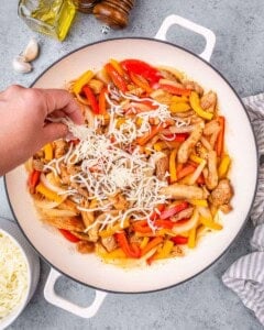 Shredded mozzarella cheese being sprinkled by hand over a mix of cooked chicken and raw peppers in a white bowl.
