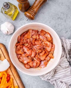 Raw sliced chicken marinating in seasoning inside a white bowl on a countertop.