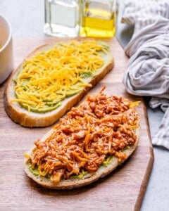 Two slices of bread, one with cheese and avocado and the other with sauce coated shredded chicken on a wooden surface.