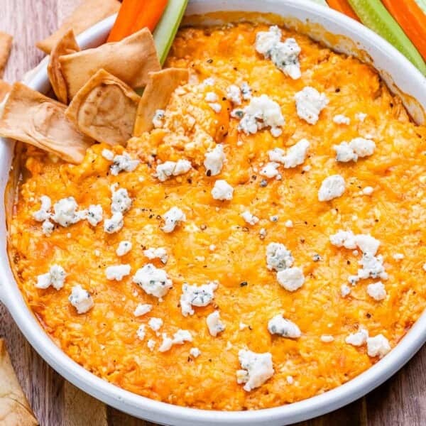 Buffalo chicken dip in a white dish with pita chips and veggies, topped with melted cheese and bits of blue cheese.