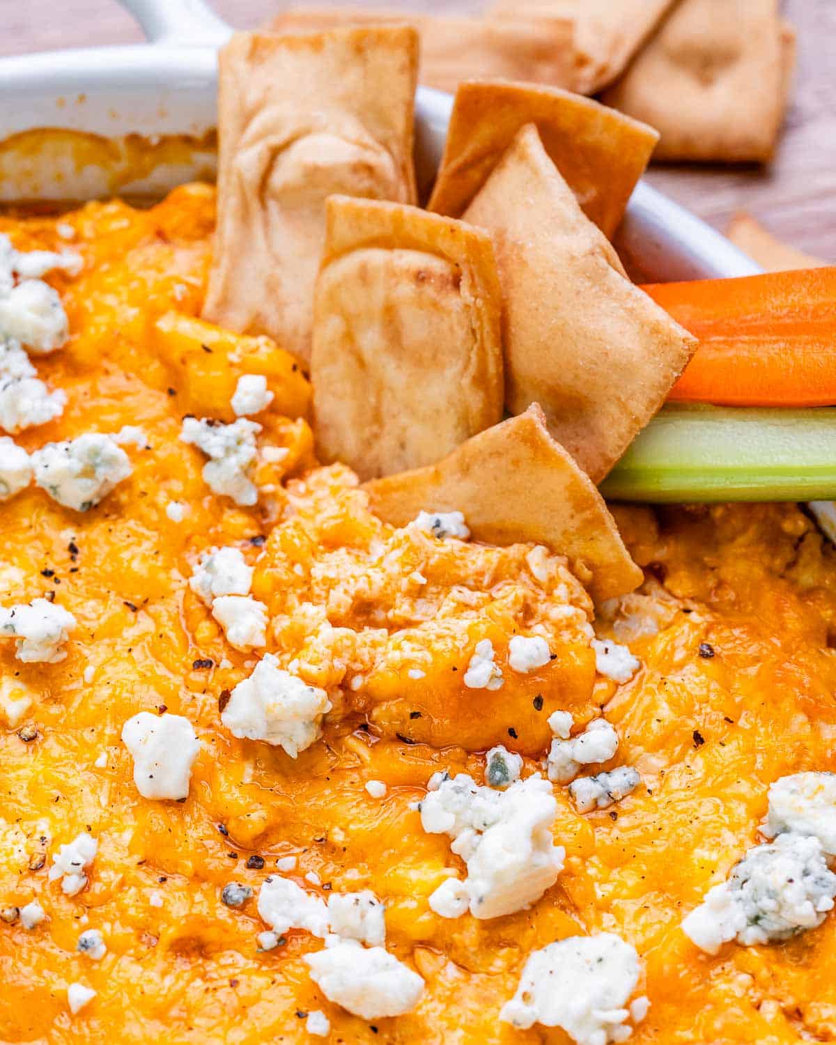 Scoop of buffalo chicken dip being lifted with a pita chip, surrounded by celery and carrots.
