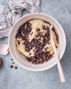 Cookie dough in a bowl with chocolate chips being folded in using a wooden spatula.