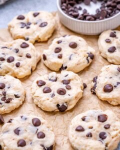 Pile of cookies on a brown surface with more chocolate chips scattered on top and around.
