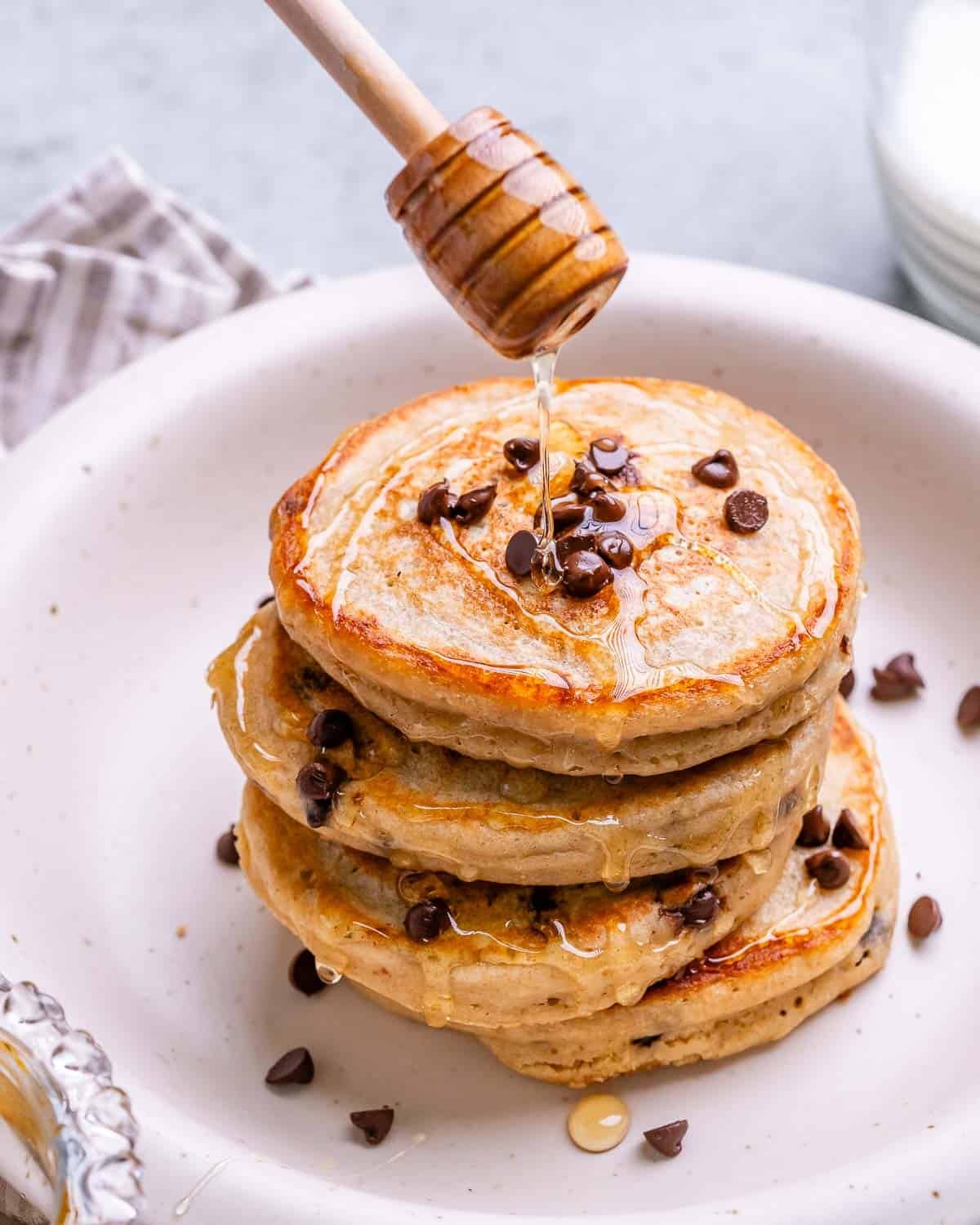Stack of Greek yogurt pancakes on a white plate drizzled with honey and sprinkled with mini chocolate chips.