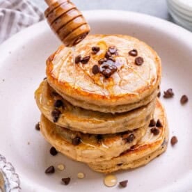 Side angle of Greek yogurt pancakes with honey being drizzled from a dipper.