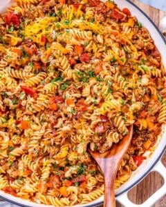 Overhead view of a large skillet filled with cheesy ground turkey pasta, garnished with fresh herbs and served with a wooden spoon.