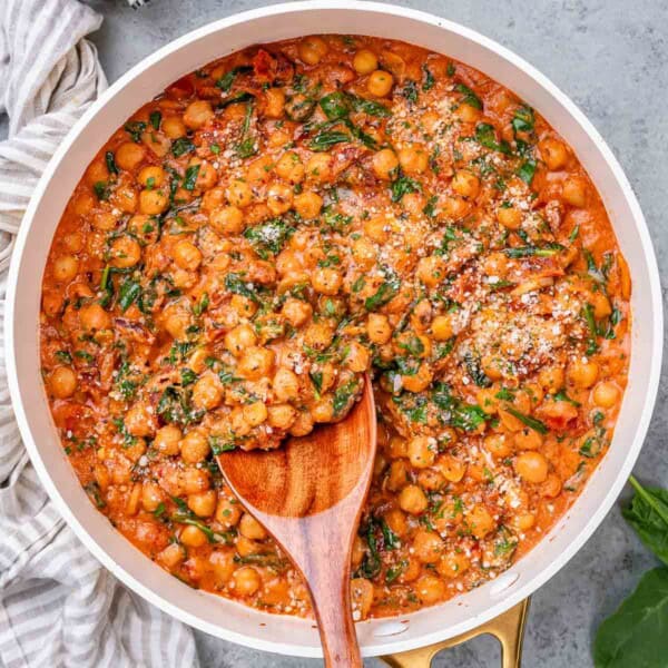 Wooden spoon stirring creamy chickpeas and spinach in a pan with a tomato-based sauce.