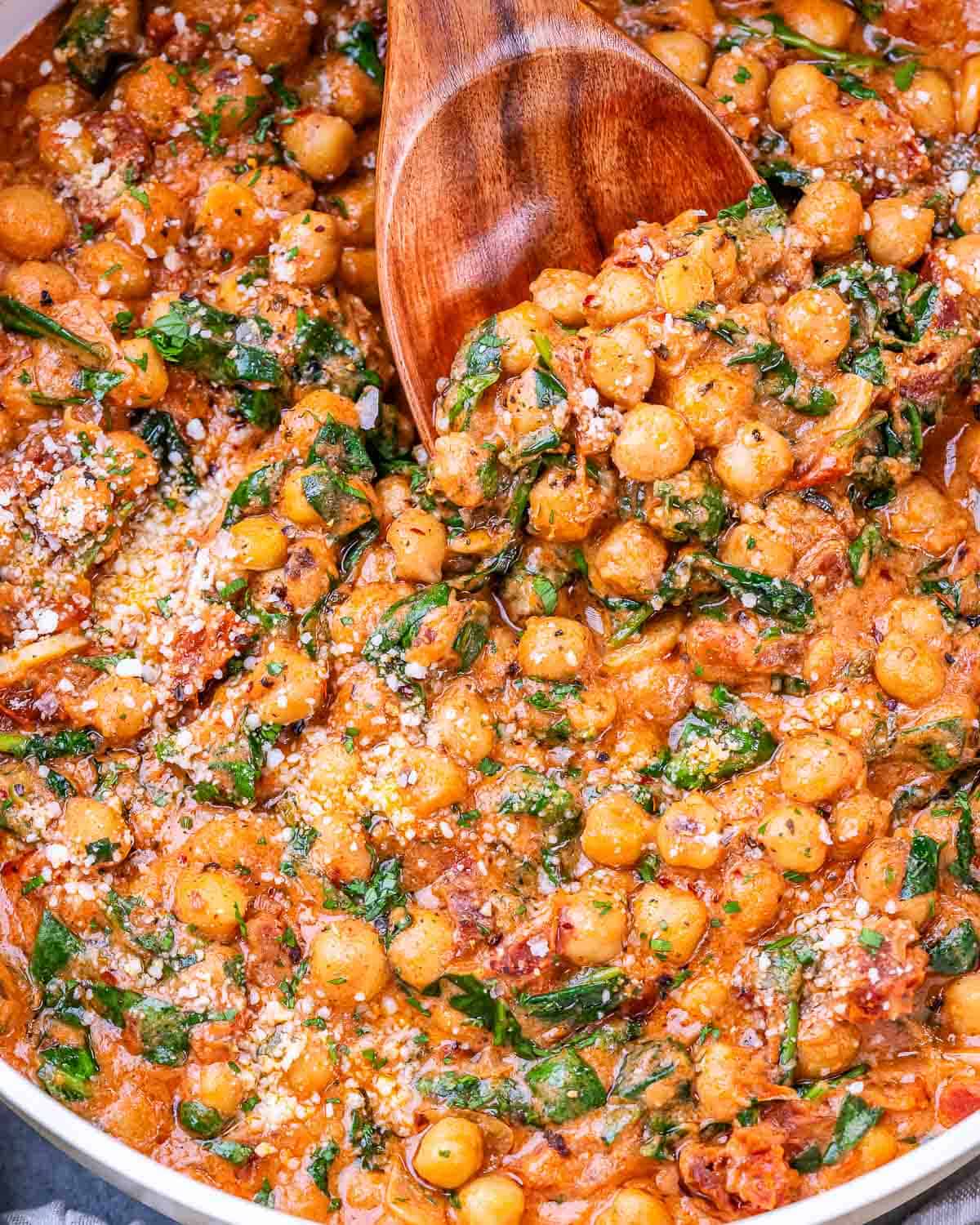 Wooden spoon scooping creamy Marry Me Chickpeas with spinach and Parmesan from a skillet.