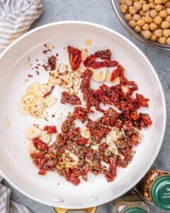 Garlic slices, chopped sun dried tomatoes, and seasonings sizzling in oil in a pan.