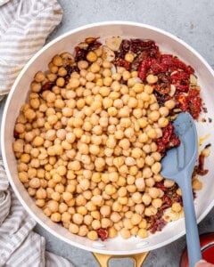 Chickpeas and chopped sun dried tomatoes being stirred together in a white skillet.