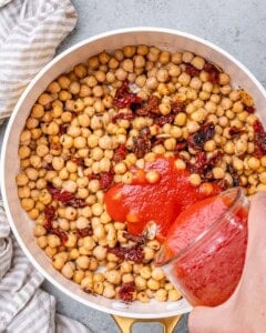Chickpeas and sundried tomatoes in a pan with tomato passata being added to the skillet before mixing.