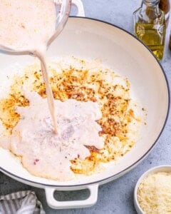 Pouring almond milk mixture into a skillet with sautéed garlic and oil.
