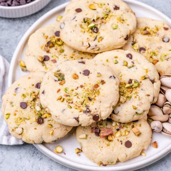 A white plate filled with pistachio chocolate chip cookies, topped with chopped pistachios and surrounded by shelled pistachios.