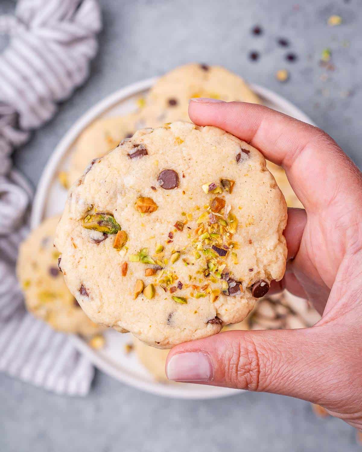 A hand holding one pistachio chocolate chip cookie, showing the soft, chewy texture with visible pistachios and chocolate chips.