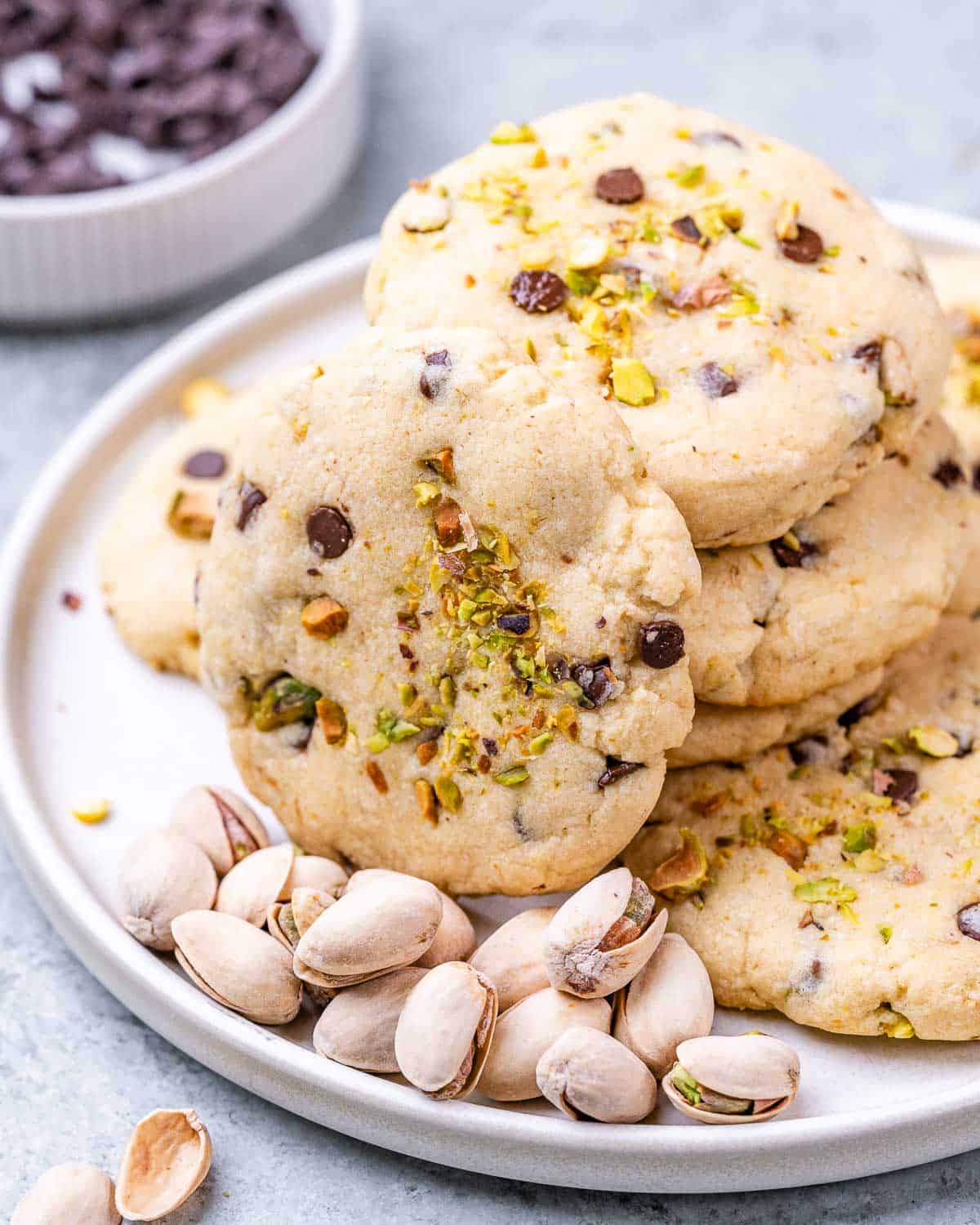 A pile of soft pistachio chocolate chip cookies on a plate with a handful of shelled pistachios scattered around the edges.