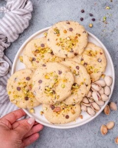 A hand holding a plate of baked pistachio chocolate chip cookies, with a linen napkin in the background and shelled pistachios nearby.