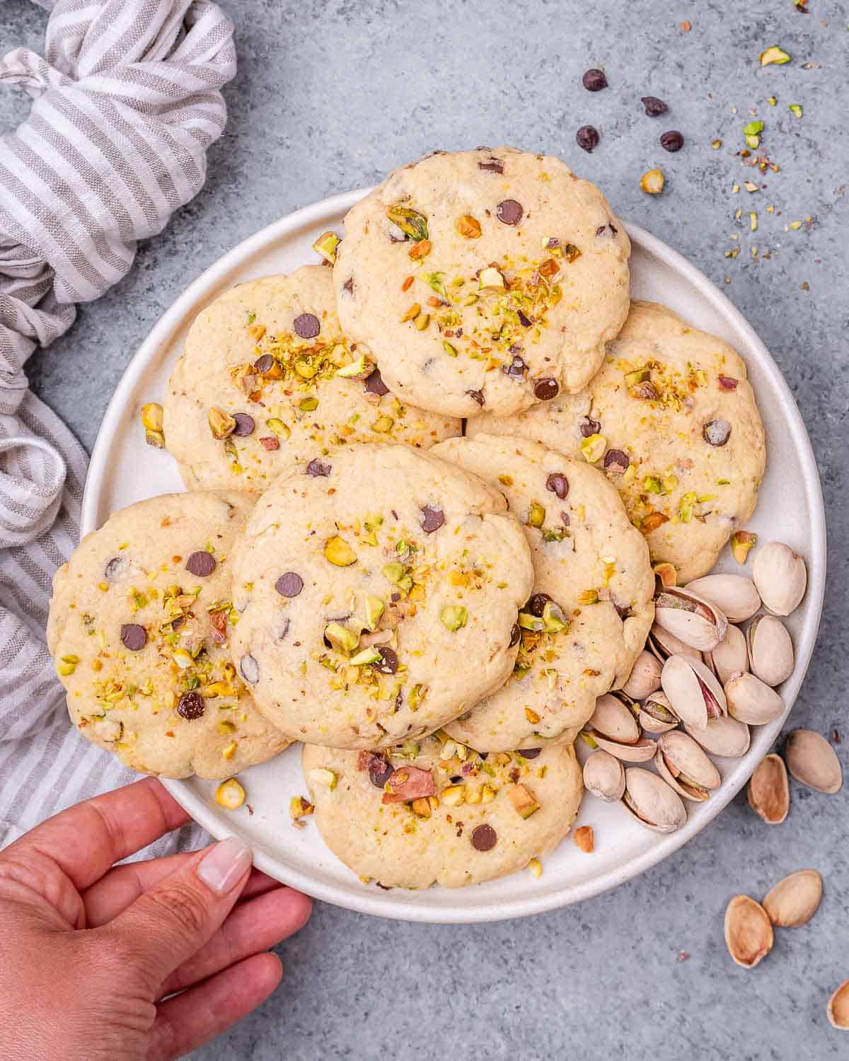 A hand holding a plate of baked pistachio chocolate chip cookies, with a linen napkin in the background and shelled pistachios nearby.