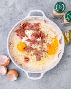 Eggs, bacon, spices, and cheese in a white baking dish before baking.